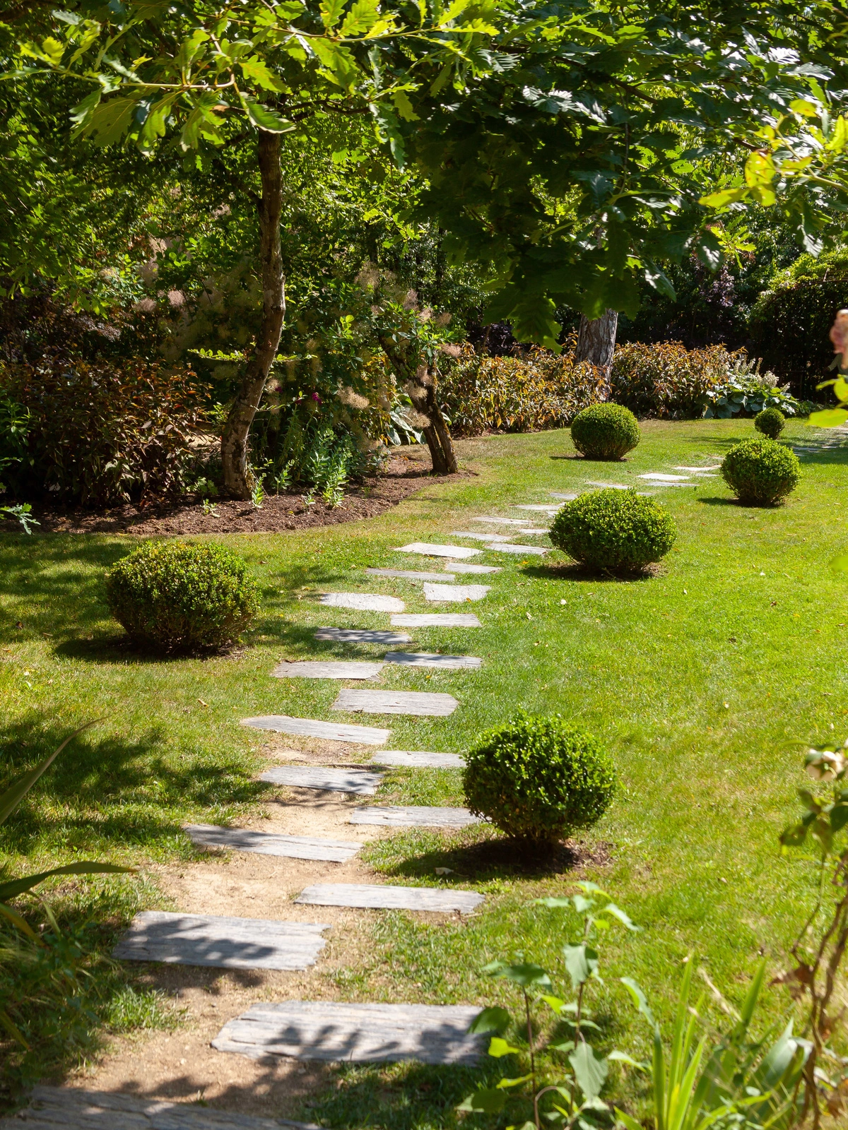 Jardin paysager avec allée en pierre, bordée de buis taillés et arbres feuillus, démontrant le travail d'un paysagiste professionnel